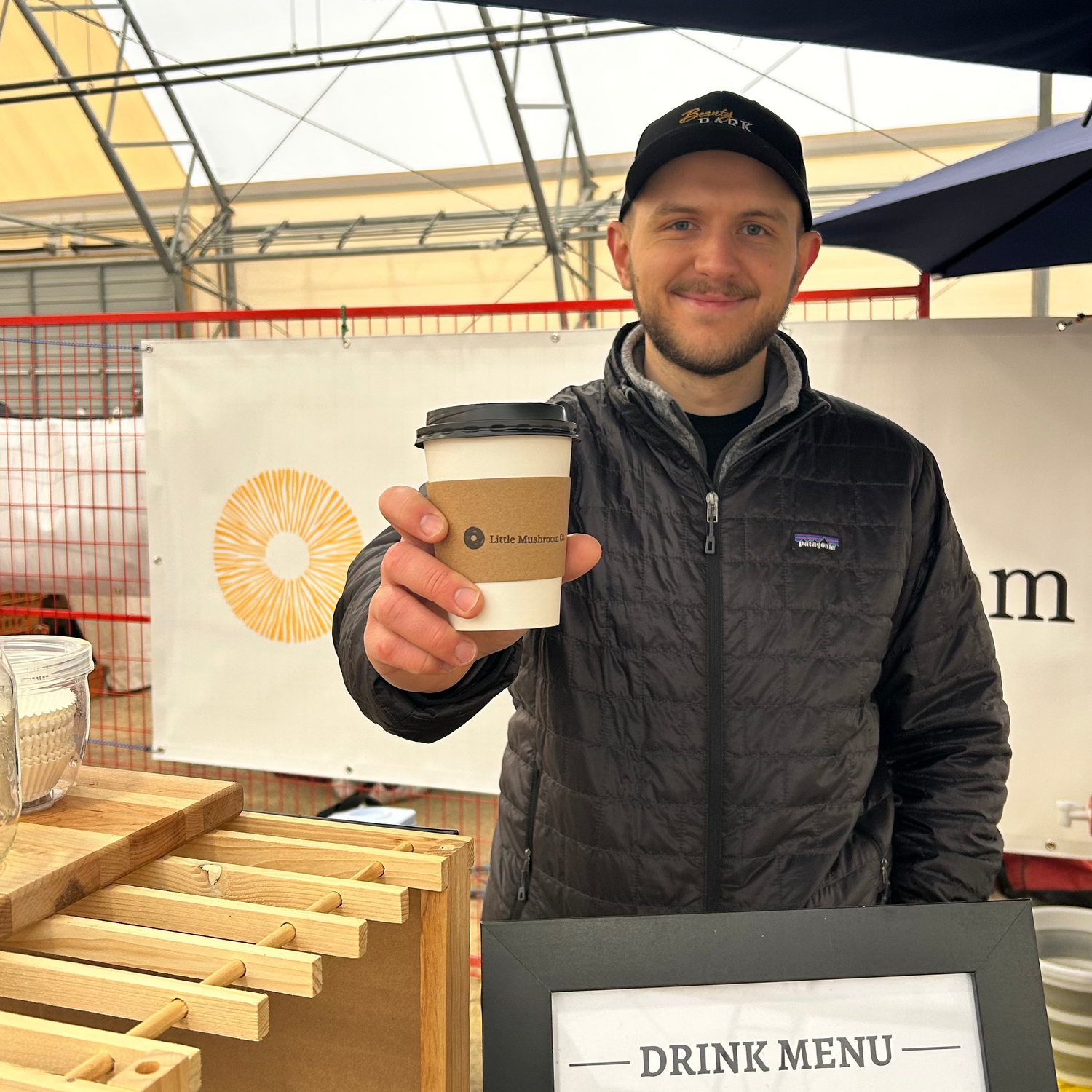 Man holding a Little Mushroom Company cup under an umbrella at a food stand with a drink menu in the foreground.